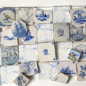 May include: A collection of antique, hand-painted ceramic tiles. The tiles feature blue and white illustrations of birds, ships, windmills, and landscapes. Some tiles are cracked or chipped, adding to their vintage charm. The tiles are arranged on a white surface.