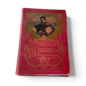 Op de afbeelding: Een vintage boek met een rode kaft en gouden letters. De kaft toont een illustratie van een man en een voertuig, met de woorden "TRAVAIL PROGRES" in sierlijk schrift. De randen van het boek vertonen tekenen van slijtage.