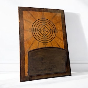 May include: A wooden board with a circular design featuring the letters "KRO" in a diamond shape. The board has a small, dark brown wooden shelf attached to the bottom.