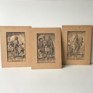 Puede incluir: Tres grabados antiguos enmarcados con grabados detallados en blanco y negro de figuras a caballo. Las impresiones están sobre un fondo beige, con signos visibles de envejecimiento y desgaste. Las imágenes representan escenas históricas.