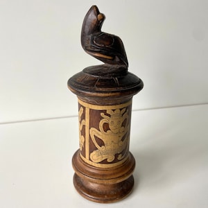 Könnte beinhalten: Dunkelbraunes dekoratives Holzobjekt mit einem geschnitzten Vogel auf einem zylindrischen Sockel. Der Sockel hat eine hellere Holzplatte mit einem geschnitzten Figurendesign. Es handelt sich wahrscheinlich um einen Dekorationsartikel oder eine kleine Skulptur.