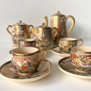 Puede incluir: Un juego de té de porcelana que incluye una tetera alta, un azucarero con tapa, una lechera y tres tazas con sus platos a juego. Cada pieza está decorada con coloridos motivos florales, pájaros y bordes dorados sobre una base de cerámica color crema.