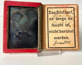 Puede incluir: Fotografía antigua en un marco rojo, con dos hombres con sombreros y trajes. La fotografía se muestra con una nota manuscrita en alemán, fechada en junio de 1904, y un soporte de vidrio transparente.
