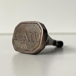 May include: Antique silver-toned wax seal with an octagonal face and a black handle. The seal's face features elegant cursive script. The seal is likely used for imprinting a design or initials on wax.