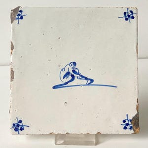 Puede incluir: Azulejo cerámico cuadrado, blanquecino, con un diseño azul pintado a mano. El diseño presenta una figura sentada, con una línea curva debajo. Acentos florales azules en cada esquina. El azulejo tiene un borde desgastado.