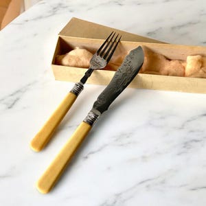 May include: A vintage silver butter knife and fork set with ivory handles in a cardboard box. The knife and fork have an ornate design.