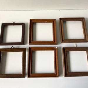 May include: A collection of six square wooden picture frames arranged in two rows of three. Each frame features a distinct brown wood grain finish, a simple border profile, and a small metal hanging hook attached to the top edge of the frame.