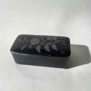 May include: A rectangular black box with a smooth, glossy finish. The lid features a silver-coloured inlay depicting a floral design with a central flower, leaves, and stems. The box has rounded corners and a solid, dark, uniform appearance.