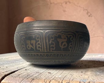 Può includere: Una ciotola tibetana in bronzo scuro con motivi e scritte incise. La ciotola ha una forma arrotondata e poggia su una superficie di legno. Un battente di legno è all'interno della ciotola.