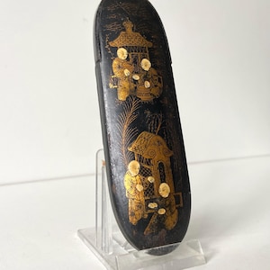 Può includere: Un astuccio nero oblungo con illustrazioni dorate di figure in un ambiente pagoda. L'astuccio è esposto in posizione verticale su un supporto di plastica trasparente. L'opera d'arte raffigura figure in abiti dorati.