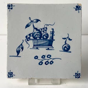May include: Square, white ceramic tile with a blue fruit basket design. The tile features a central basket filled with fruit, surrounded by floral and leaf motifs. The corners have small floral accents.