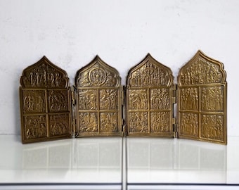 May include: A vintage, brass-colored, folding icon with intricate relief carvings. The icon is divided into four panels, each with detailed religious scenes. The top of each panel has an arched design. The icon is displayed on a white surface.