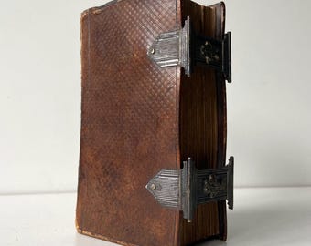 May include: A vintage, leather-bound book with a textured brown cover and metal clasps. The book's pages are aged, and the metal clasps are ornate. The book is standing upright, showcasing its aged appearance and craftsmanship.