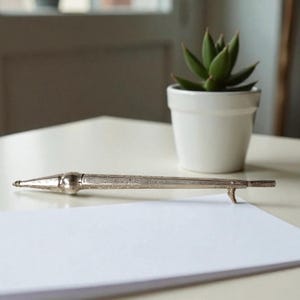 Puede incluir: Un lápiz antiguo plateado con detalles grabados descansa sobre una superficie blanca, junto a una pequeña suculenta en una maceta blanca. El lápiz tiene una punta afilada y un gancho en el extremo. Un papel blanco en blanco está en primer plano.