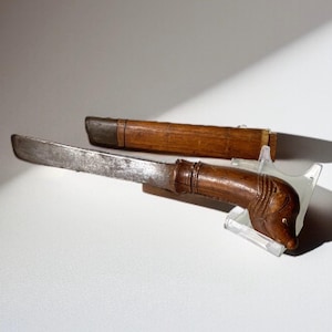 Puede incluir: Un cuchillo antiguo con un mango de madera tallado en forma de cabeza de animal. La hoja es plateada y el mango es de color marrón oscuro. Una vaina de madera está detrás del cuchillo.