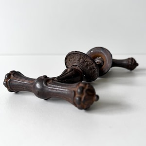 May include: A pair of dark brown, weathered metal door handles featuring an ornate, textured design with rounded ends and a circular mounting plate. The hardware displays a rustic, aged finish with visible oxidation and intricate detailing.