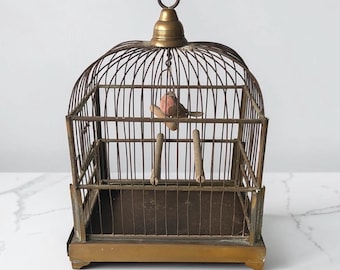 Op de afbeelding: Een antieke vogelkooi met een kleine, decoratieve vogel erin. De kooi is gemaakt van metaal met een messing afwerking en heeft een gewelfde bovenkant met een ring. De vogel is roze en bruin, zittend op een houten deuvel. De kooi is een vintage decoratief item.