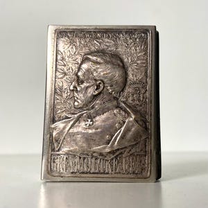 Pode incluir: Placa retangular de metal prateado com um perfil detalhado de um homem em uniforme militar. A placa é adornada com texto e detalhes de folhas de louro. As palavras "GRAF MOLTKE" são visíveis.