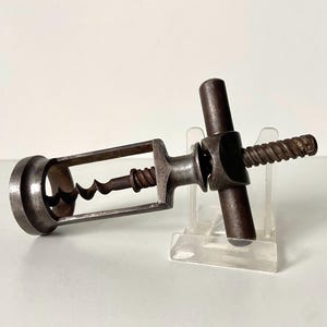 May include: A vintage metal corkscrew with a dark, aged finish. The corkscrew has a spiral worm, a handle, and a frame. The design suggests a classic style, suitable for wine enthusiasts or collectors. The corkscrew is approximately 15 cm in length.