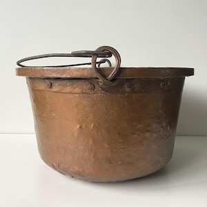 May include: A copper cauldron with a rounded base and a sturdy metal handle. The pot has a wide rim and is riveted for durability. The aged copper has a warm, reddish-brown hue, suggesting it is an antique or vintage piece.