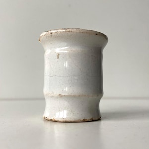 May include: A small, off-white ceramic vase with a cylindrical shape. The vase has a slightly flared rim and a base that tapers inward. The surface shows subtle cracks and brown discoloration, suggesting age or a rustic aesthetic.