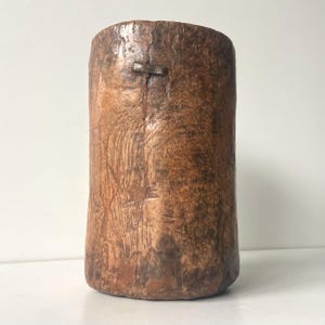 Könnte beinhalten: Ein zylindrischer, rustikaler Holzbehälter mit natürlichem, gealtertem Aussehen. Die Holzmaserung ist sichtbar, und die Farbe reicht von hell- bis dunkelbraun. Eine Metallklammer sichert einen Riss an der Seite. Der Behälter hat einen glatten, abgerundeten Boden.