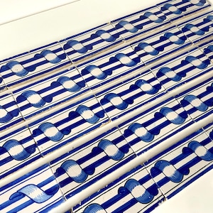Op de afbeelding: Een verzameling rechthoekige keramische tegels met een herhalend patroon. Elke tegel heeft een witte achtergrond met blauwe strepen en een centraal ontwerp van een blauw-witte gestileerde vorm. De tegels zijn in rijen gerangschikt.