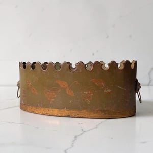 May include: An oval metal container with a scalloped top edge and a muted olive green finish. The exterior features a painted copper-colored grape and leaf motif. It includes small metal ring handles on each side and a textured copper base.