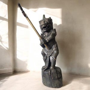 May include: A detailed, dark-toned statue of a mythical figure holding a spear. The figure has an expressive face and is adorned with intricate details. The spear has a gold-coloured shaft and a dark tip. The statue is mounted on a dark base.