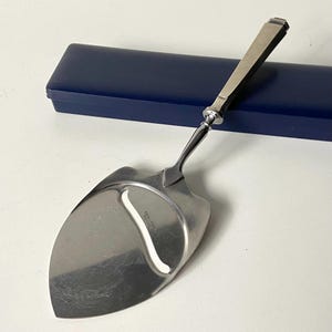May include: A silver cheese knife with a wooden handle and a blue velvet box. The knife has a serrated edge and a small hole in the blade. The box is closed.