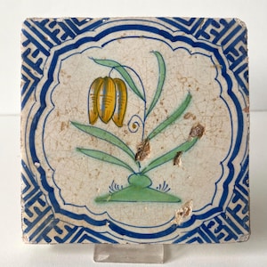 May include: A square ceramic tile with a white base and a blue geometric border. The center features a hand-painted illustration of a yellow flower with green leaves and a green base. The surface shows visible crackling throughout the glaze.