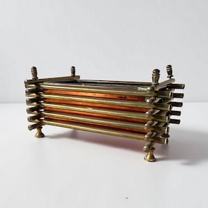 Puede incluir: Un recipiente decorativo rectangular con interior de cobre y exterior de latón. El recipiente está construido con varillas horizontales de latón y soportes verticales, y descansa sobre cuatro pequeños pies de latón. El recipiente tiene una estética vintage.
