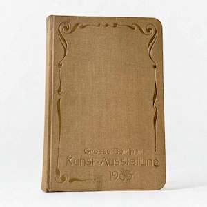 May include: A vintage book with a tan cover and decorative embossed border. The cover features the text "Grosse Berliner Kunst-Ausstellung 1905" in a serif font. The book has a rectangular shape and a slightly worn appearance.