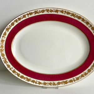 Può includere: Un piatto da portata ovale con sfondo bianco e una larga fascia rosso cremisi intorno al bordo. La fascia è decorata con un motivo a vite dorato.