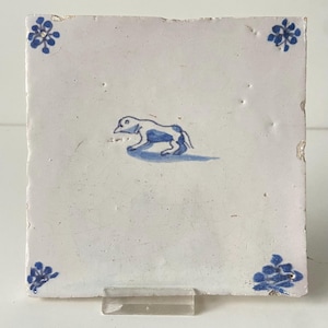 May include: Square white ceramic tile with a blue and white illustration of a dog. The tile has blue floral corner accents. The dog is depicted in a simple, stylized manner, with blue shading and outlines.