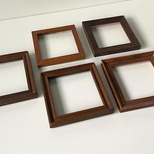 May include: A collection of five square wooden frames in varying shades of brown, ranging from light tan to dark espresso. Each frame features a simple, flat-profile design with mitred corners, arranged in a cluster on a plain white surface.