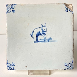 May include: Square ceramic tile with a hand-painted design of a rabbit in blue and white. The rabbit is depicted in a playful pose, with its ears perked up. Decorative blue floral corner accents complete the design.
