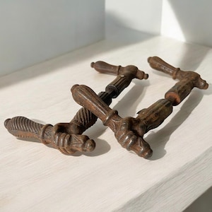 Peut inclure: Quatre poignées de porte antiques en bronze avec des motifs ornés et texturés. Les poignées sont disposées sur une surface blanche, mettant en valeur leur aspect vieilli et patiné. Les poignées ont une forme en T.