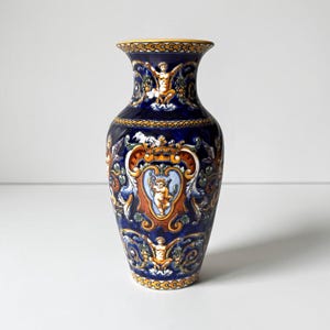 Może przedstawiać: Ceramiczny wazon o głębokim niebieskim kolorze podstawy, ozdobiony ręcznie malowanymi wzorami w kolorach złotym, białym i pomarańczowym. Powierzchnia przedstawia ozdobne zawijasy, motywy kwiatowe oraz centralne kartusze z postaciami cherubinów.