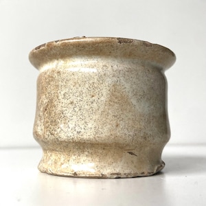 May include: A small, antique, off-white ceramic pot with a speckled surface. The pot has a wide, slightly flared rim and a rounded base. The exterior shows signs of age and wear, with a distressed appearance.
