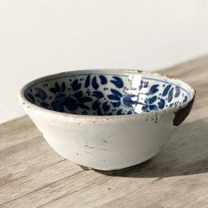 Może przedstawiać: Mała biała ceramiczna miska z niebiesko-białym wzorem kwiatowym. Miska ma odprysk na brzegu i nosi ślady zużycia.