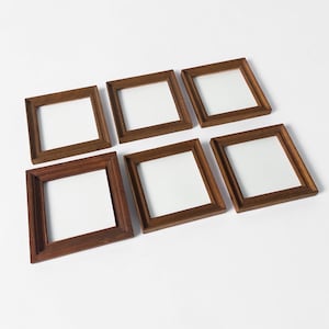 Puede incluir: Seis marcos de fotos cuadrados de madera con acabado marrón oscuro. Cada marco tiene un interior blanco, creando contraste con la madera. Los marcos están dispuestos sobre una superficie blanca.