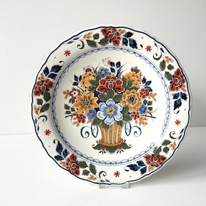 May include: A round ceramic plate with a scalloped edge featuring a central hand-painted floral arrangement in a basket. The design includes yellow, blue, and red flowers with green leaves, surrounded by a blue lattice border and floral motifs.