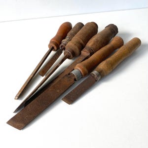 May include: A collection of five vintage hand tools featuring weathered brown wooden handles and oxidized metal shafts. The set includes various files and chisels with distinct textures, showing signs of age and use with dark metal surfaces.