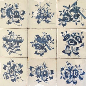 Könnte beinhalten: Neun quadratische Fliesen mit handgemalten blauen Blumen- und Vogelmotiven auf cremefarbenem Hintergrund. Jede Fliese zeigt eine einzigartige Anordnung von Blumen, Blättern und Vögeln, die ein zusammenhängendes, aber abwechslungsreiches Muster bilden. Die Fliesen sind wahrscheinlich aus Keramik.