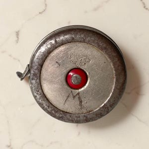 May include: A vintage circular metal measuring tape with a weathered silver finish and a bright red centre button. The casing features the engraved text E.M.D. DURLACH and a hand-etched letter M. A small metal tab protrudes from the side of the housing.