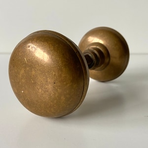 Puede incluir: Un pomo de puerta de latón vintage con forma redonda y ligeramente abovedada. El pomo tiene una superficie lisa y desgastada y un borde ligeramente elevado.