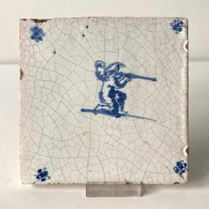 Op de afbeelding: Een vierkante keramische tegel met een wit geglazuurd oppervlak en een blauwe, handgeschilderde afbeelding van een knielende figuur die op een lang voorwerp richt. De tegel heeft een zichtbare craquelé textuur en kleine blauwe hoekmotieven.