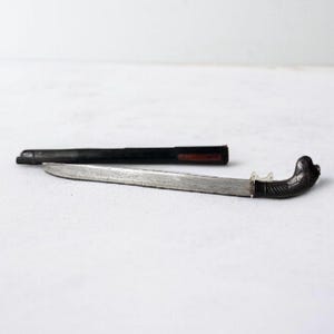 Könnte beinhalten: Ein Messer mit silberner Klinge, einem geschwungenen, verzierten Griff und einer schwarzen Scheide. Der Griff hat ein dunkles, strukturiertes Aussehen. Die Scheide ist schwarz mit einem roten Akzent in der Nähe der Öffnung. Das Messer liegt auf einer weißen Oberfläche.