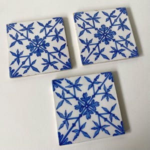 May include: Three square ceramic tiles featuring a white base with a repeating blue floral and geometric pattern. Each tile displays a central stylized flower motif with radiating lines extending toward the corners, creating a symmetrical design.
